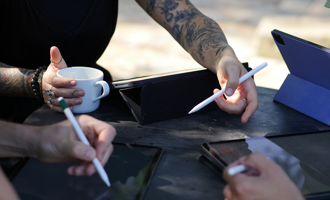 Two people sketching on iPads with Apple Pencils at an outdoor table, one holding a coffee mug